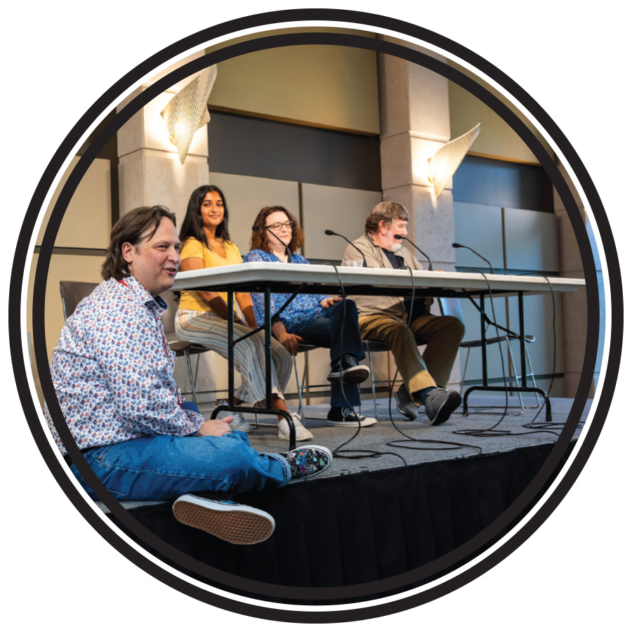 Three people sit at a table on a stage in front of microphones and one sits on the edge of the stage