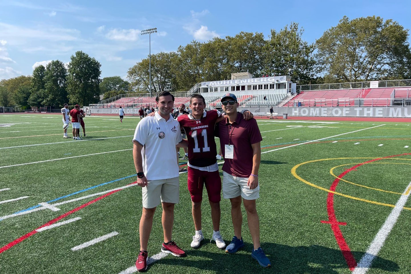 Three people smile at the camera while standing on a football field